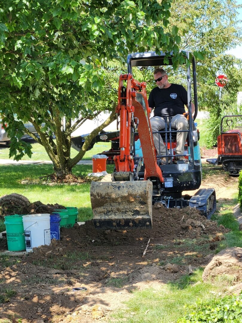 All-Pro Plumbing technician operating an excavator for traditional drain repair and perimeter drain or sewer line excavation at a residential site in Kokomo, IN