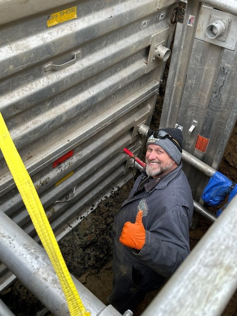 All-Pro Plumbing technician giving a thumbs up from inside a safe trench box during a deep drain repair in a Kokomo, IN alleyway