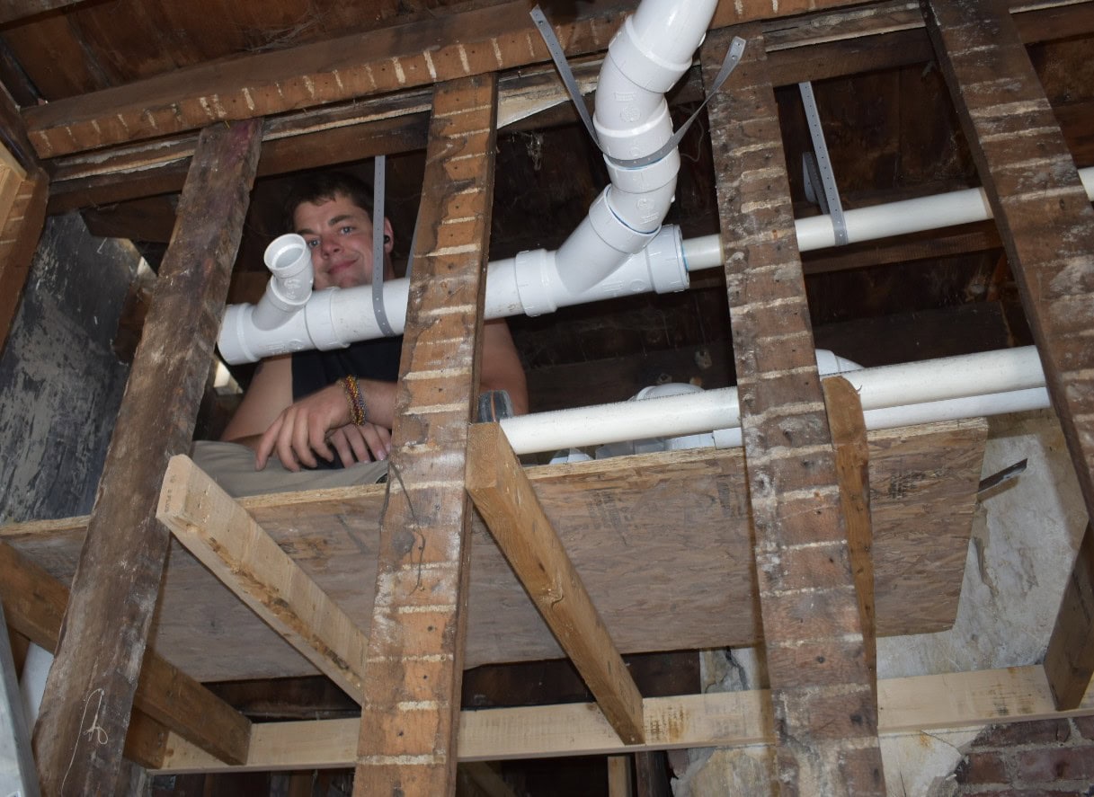 All-Pro Plumbing technician smiling while installing new PVC drain pipes in a ceiling as part of the Bridges Outreach Program in Kokomo, IN