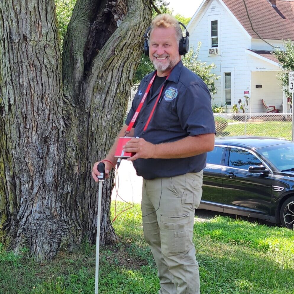 All-Pro Plumbing technician smiling and using acoustic listening device for outdoor leak detection near a tree in Kokomo, IN