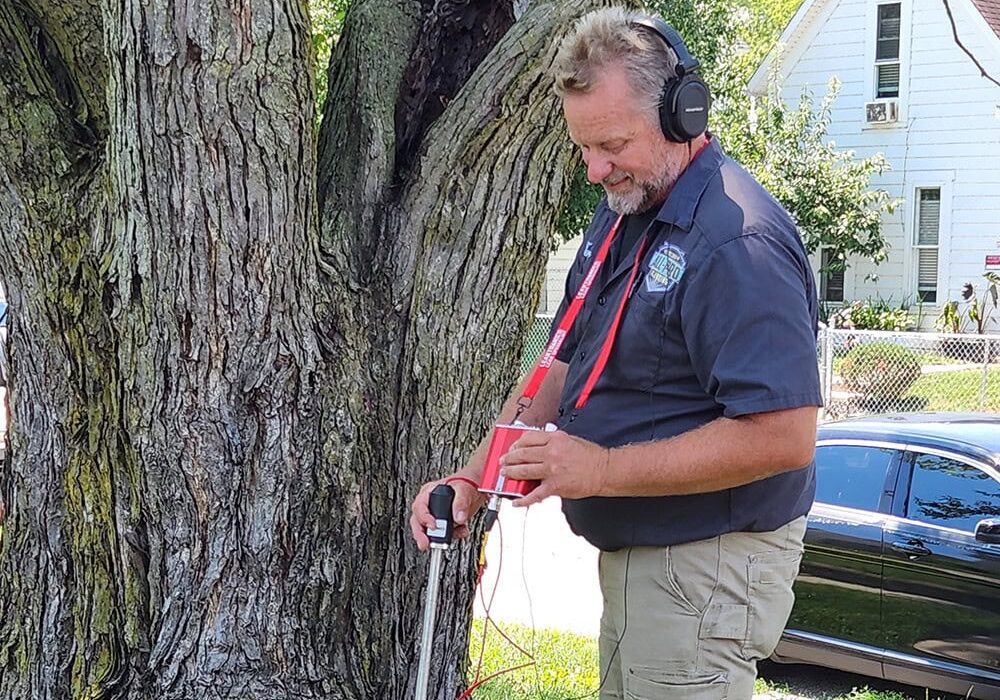 3rec Leak Detection Yard High Water Bill Locate All-Pro Plumbing technician smiling and using acoustic listening device for outdoor leak detection near a tree in Kokomo, IN
