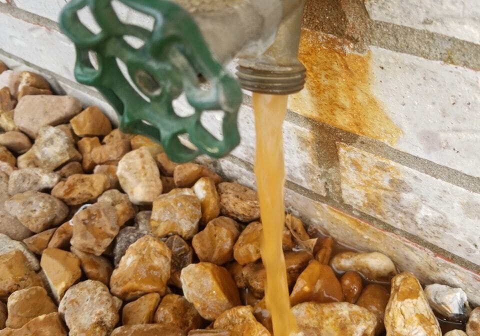 47 Water Treatment Conditioning Iron Well Water Outside Hydrant Spigot Faucet Orange Staining Outdoor spigot with orange-stained water flowing onto rocks, indicating high iron content in well water, requiring water treatment and conditioning by All-Pro Plumbing in Kokomo, IN