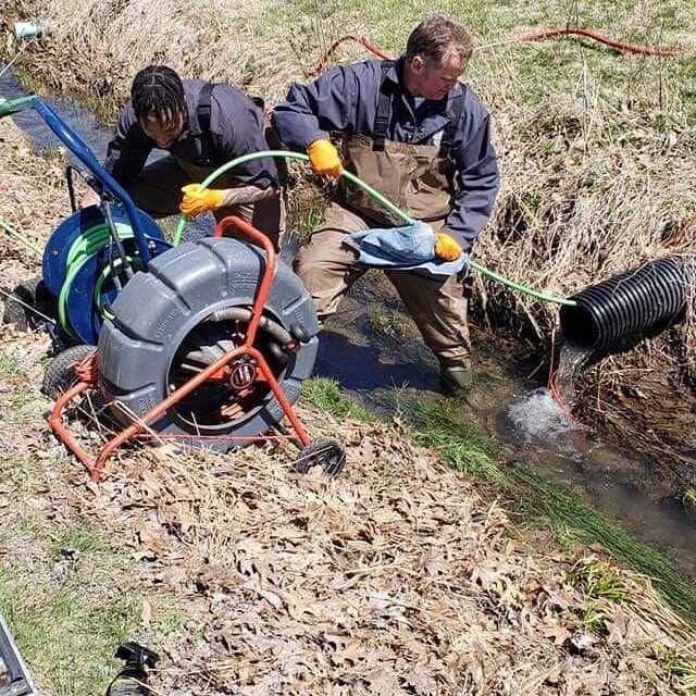 Two All-Pro Plumbing technicians performing hydro-jetting drain cleaning on an outdoor perimeter drain in a ditch using specialized equipment in Kokomo, IN