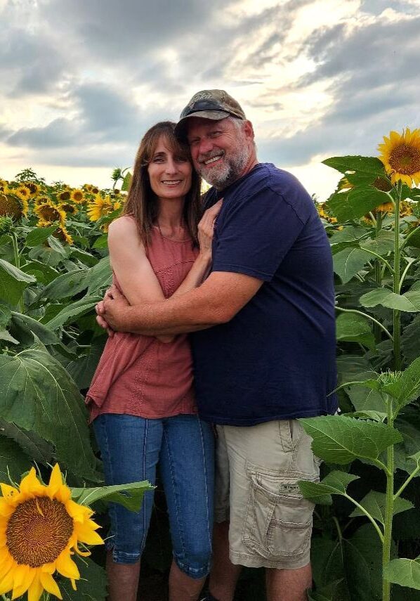Kim and Terry Tracy, owners of All-Pro Plumbing, smiling and embracing in a beautiful sunflower field in Kokomo, IN, representing the family-owned business