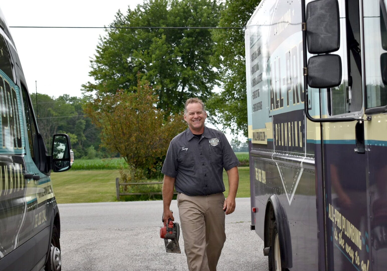 Owner Terry Tracy of All-Pro Plumbing smiling while walking between two branded company fleet vans in Kokomo, IN