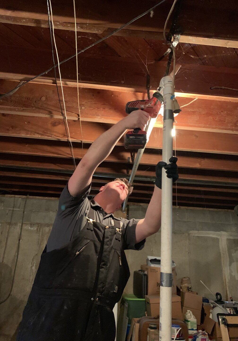 77 Basement Repipe Expert Sump Pump Drain Check Valve Flooding All-Pro Plumbing expert repiping a sump pump drain line in a basement with a power drill, to prevent flooding in a Kokomo, IN home