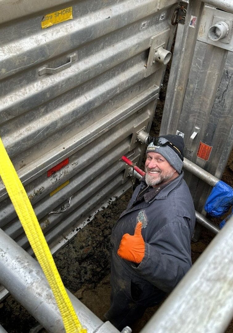 81 Alleyway Street Cut Trench Boxes Safety First Deep Drain Repair All-Pro Plumbing technician giving a thumbs up from inside a safe trench box during a deep drain repair in a Kokomo, IN alleyway